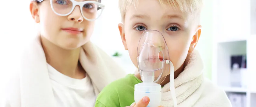 Chłopiec trzymający inhalator dłoni i przeprowadza inhalacja. Za chłopcem stoi dziewczynka.