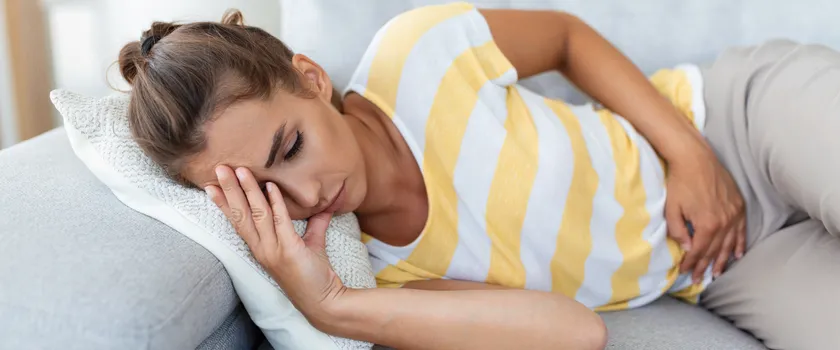 Kobieta w bluzce w paski leży na łóżku i trzyma się swój brzuch.