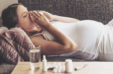 Kobieta leżąca na łóżku i wydmuchująca nos. Kobieta jest w ciąży i ubrana na biało.