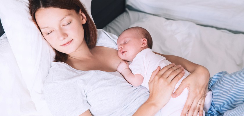 Małe dziecko śpi w rękach matki. Kobieta leży na łóżku z zamkniętymi oczami.