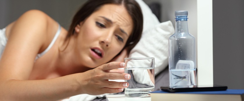 Spragniona kobieta w łóżku sięga po szklankę wody z szafki nocnej.