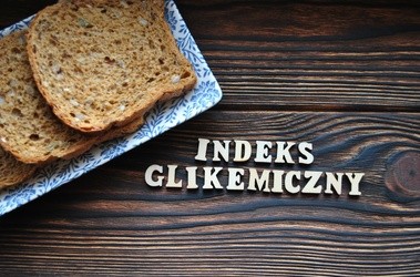 Kawałki chleba położone na talerzu, który jest na drewnianym blacie. Obok ułożony z literek napisz "indeks glikemiczny"