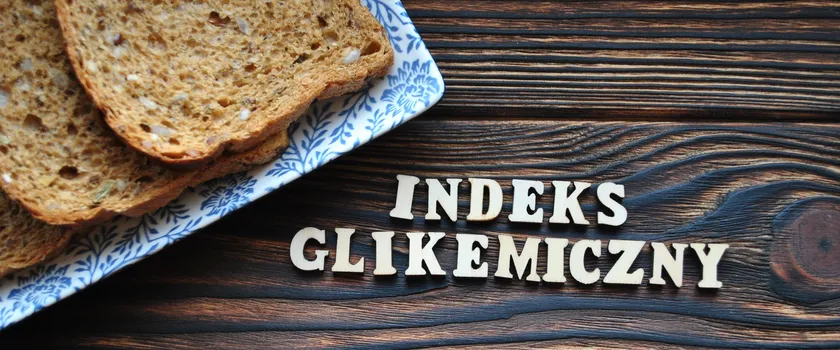 Kawałki chleba położone na talerzu, który jest na drewnianym blacie. Obok ułożony z literek napisz "indeks glikemiczny"