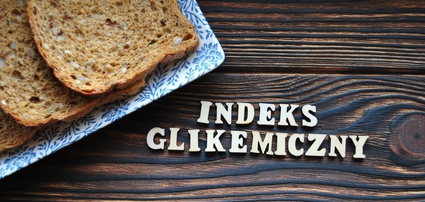 Kawałki chleba położone na talerzu, który jest na drewnianym blacie. Obok ułożony z literek napisz "indeks glikemiczny"