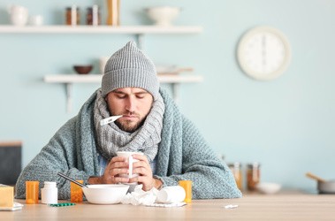 Mężczyzna ubrany w sweter, szalik oraz czapkę. Mężczyzna trzymający termometr w ustach oraz kubek z ciepłym napojem. Na stole leżą lekarstwa.