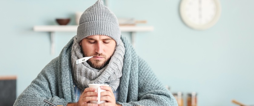 Mężczyzna ubrany w sweter, szalik oraz czapkę. Mężczyzna trzymający termometr w ustach oraz kubek z ciepłym napojem. Na stole leżą lekarstwa.