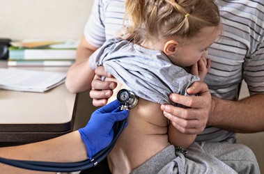 Mała dziewczynka z ojcem w gabinecie lekarskim. Lekarz bada dziecko, słucha fonendoskopem płuca dziewczynki.