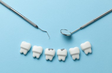Sześć białych zębów na niebieskim tle. Dwa z nich zawiera ślady próchnicy. Obok zębów narzędzia dentystyczne.