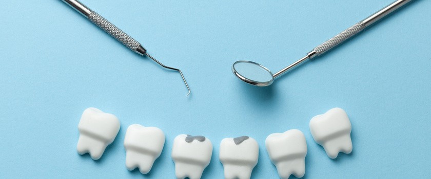 Sześć białych zębów na niebieskim tle. Dwa z nich zawiera ślady próchnicy. Obok zębów narzędzia dentystyczne.