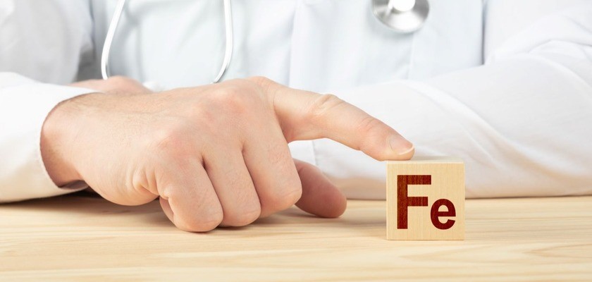Nadmiar żelaza: dłoń mężczyzny w kitlu wskazującego na drewniany klocek z literami FE (symbol żelaza). 