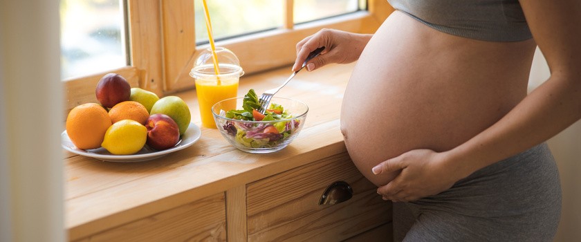 Pani w zaawansowanej ciąży je zdrową żywność w domu. Na blacie stoją owoce, zdrowa sałatka z warzyw oraz sok pomarańczowy.