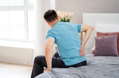 Mężczyzna siedzący na łóżku i trzymający się za dolną cześć pleców z powodu bólu nerek.