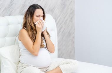 Kobieta w ciąży wydmuchuje nos siedząc na łóżku.