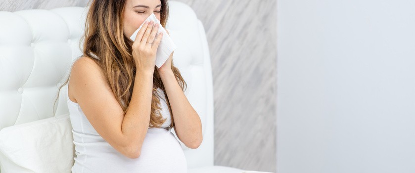 Kobieta w ciąży wydmuchuje nos siedząc na łóżku.