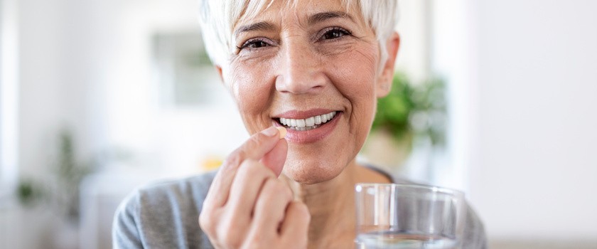 Starsza kobieta trzymająca w jednej dłoni tabletkę, a w drugiej szklankę wody.