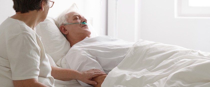 starszy człowiek w śpiączce leżący na szpitalnym łóżku i jego żona siedzi obok niego