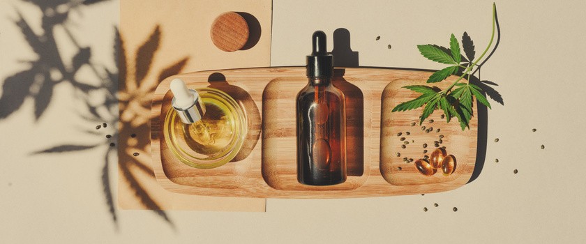 Olej CBD, nalewka z pipetą oraz tabletka na drewnianej desce na beżowym tle z liśćmi konopi.