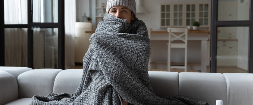 Przeziębiona kobieta w czapce zawinięta w koc, siedząca na łóżku w salonie.