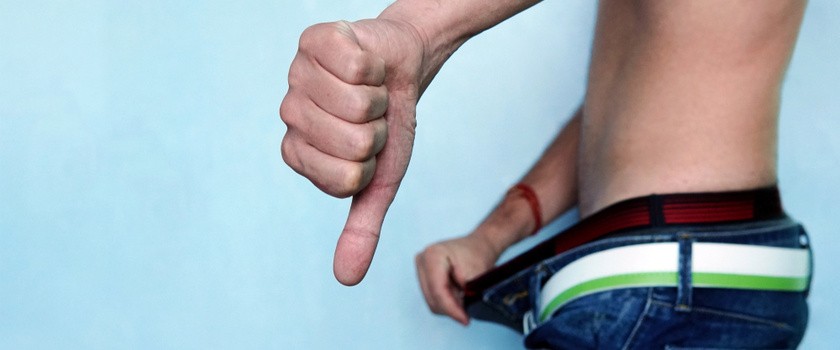 mężczyzna bez koszulki w jeansach z problemem niepełnego wzwodu na tle niebieskiej ściany pokazuje palec w dół