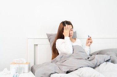 Młoda kobieta z przeziębieniem lub COVID leży w łóżku w białej pościeli. Mierzy temperaturę i dotyka czoła
