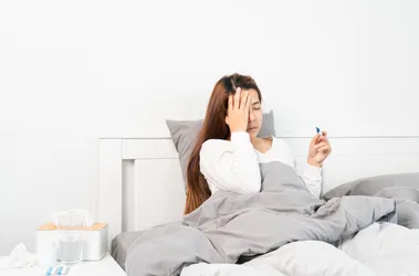 Młoda kobieta z przeziębieniem lub COVID leży w łóżku w białej pościeli. Mierzy temperaturę i dotyka czoła