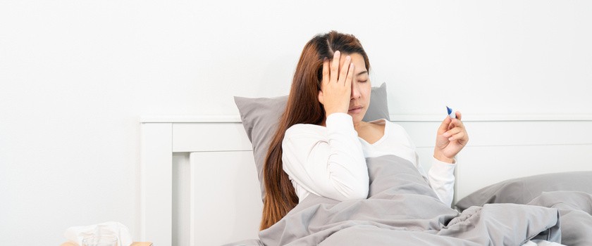 Młoda kobieta z przeziębieniem lub COVID leży w łóżku w białej pościeli. Mierzy temperaturę i dotyka czoła