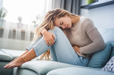 Młoda kobieta cierpi na silny ból brzucha, siedząc na kanapie w domu.