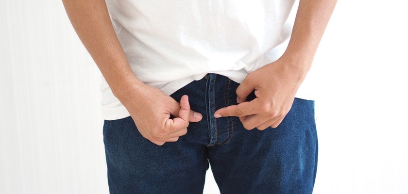 mężczyzna na białym tle ubrany w białą bluzkę i jeansy wskazuje palcami na genitalia