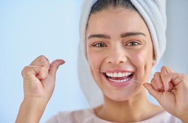 Młoda uśmiechnięta kobieta podczas nitkowania zębów. Kobieta ma zawinięty ręcznik na głowie.