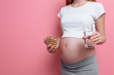Kobieta w ciąży na różowym tle trzymająca tabletki z żelazem. Przyjmowanie żelaza w ciąży