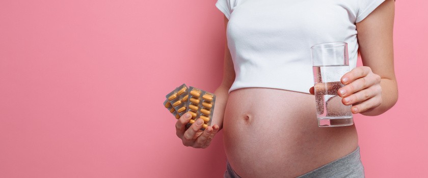 Kobieta w ciąży na różowym tle trzymająca tabletki z żelazem. Przyjmowanie żelaza w ciąży