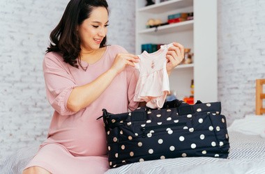 Kobieta w ciąży siedzi na łóżku i pakuje rzeczy do torby do porodu.