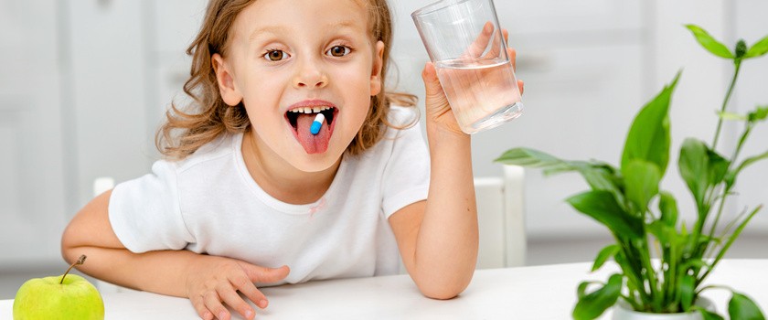 Uśmiechnięta dziewczynka mająca na języku tabletkę, a w rączce szklankę wody.