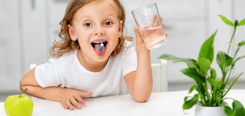 Uśmiechnięta dziewczynka mająca na języku tabletkę, a w rączce szklankę wody.