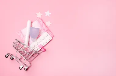 Wózek sklepowy wraz z akcesoriami wykorzystywanymi podczas miesiączki: tamponami, podpaskami, tabletkami oraz kubeczkiem menstruacyjnym.
