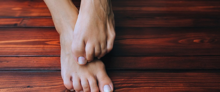 Kobiece stopy na tle brązowej drewnianej podłogi z bliska. Swędzenie i drapanie z powodu grzybicy.