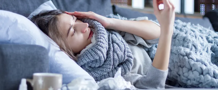 Kobieta leżąca na łóżku otulona w koc i mierząca temperaturę. Obok leżą leki, cytryna oraz kubek.