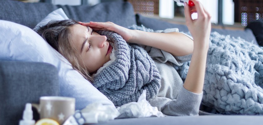 Kobieta leżąca na łóżku otulona w koc i mierząca temperaturę. Obok leżą leki, cytryna oraz kubek.