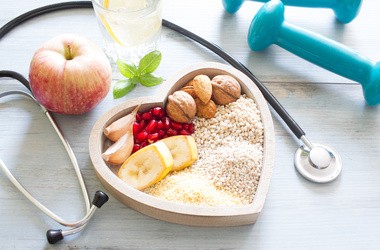 Drewniane pojemnik w kształcie serce wypełniony zdrową żywnością. Obok szklanka wody, hantle, jabłko oraz stetoskop.