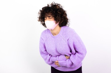 Młoda kobieta w maseczce z biegunką jako objaw koronawirusa. Kobieta w fioletowym swetrze trzyma się za bolący brzuch na białej ścianie