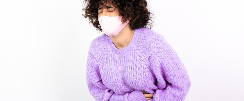 Młoda kobieta w maseczce z biegunką jako objaw koronawirusa. Kobieta w fioletowym swetrze trzyma się za bolący brzuch na białej ścianie